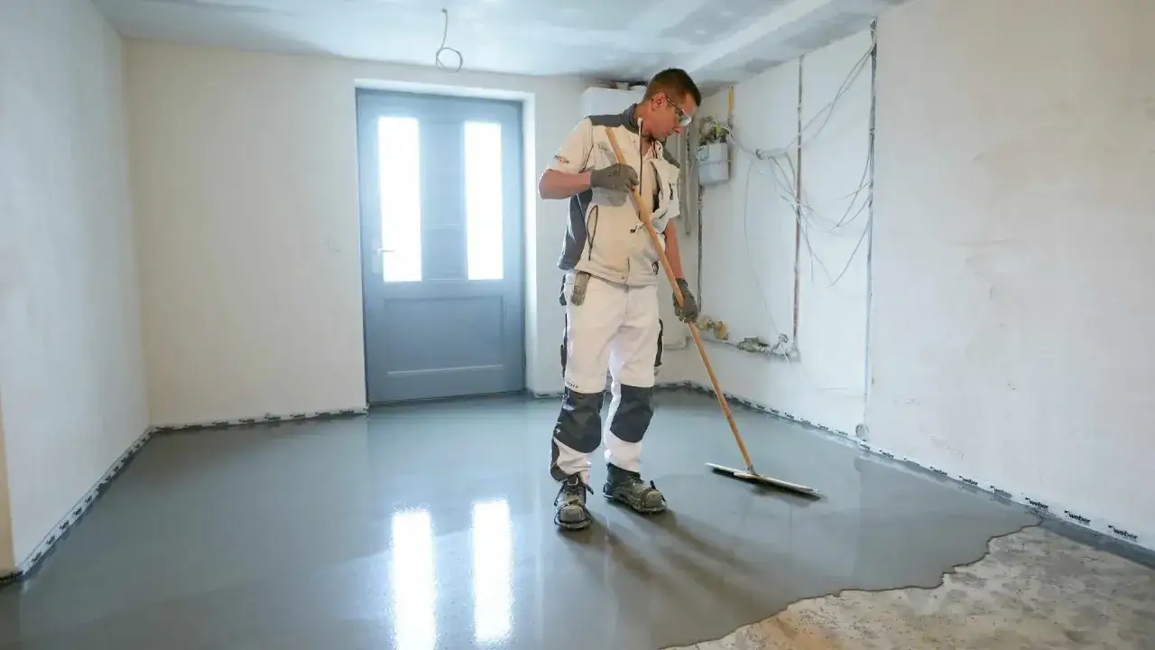 Handwerker verteilt Estrich auf der Bodenplatte eines Altbaus. Die glatte Oberfläche wird mit einem Abzieher bearbeitet, um Risse zu vermeiden.