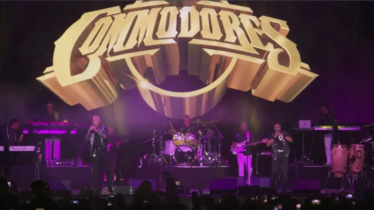Koncert zespołu Commodores, gdzie każdy dźwięk jest **easy like Sunday morning**. Wokalista śpiewa, a zespół gra na instrumentach pod złotym logo.