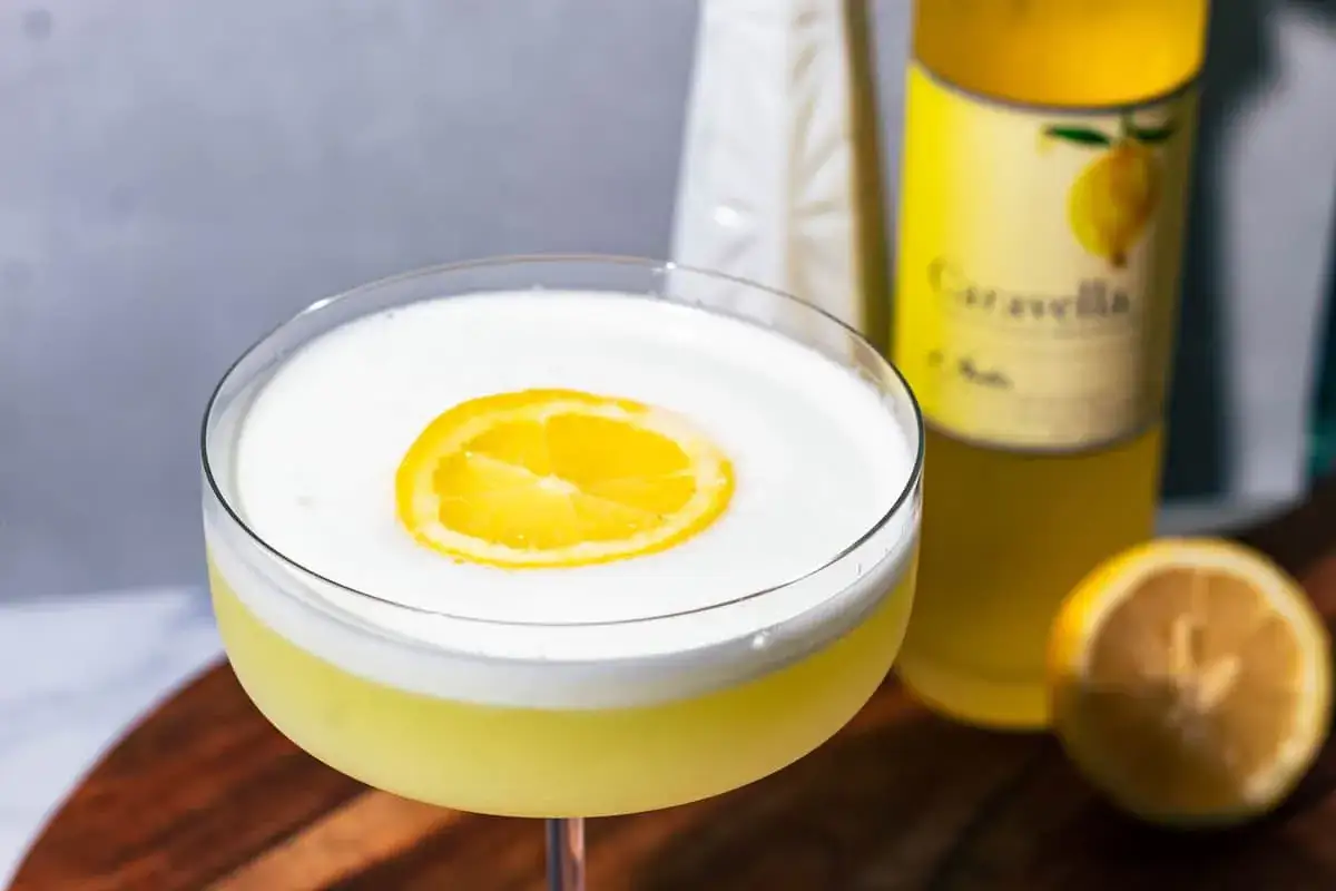 En krämig limoncello sour med en citronskiva på toppen, bredvid en flaska Caravella.