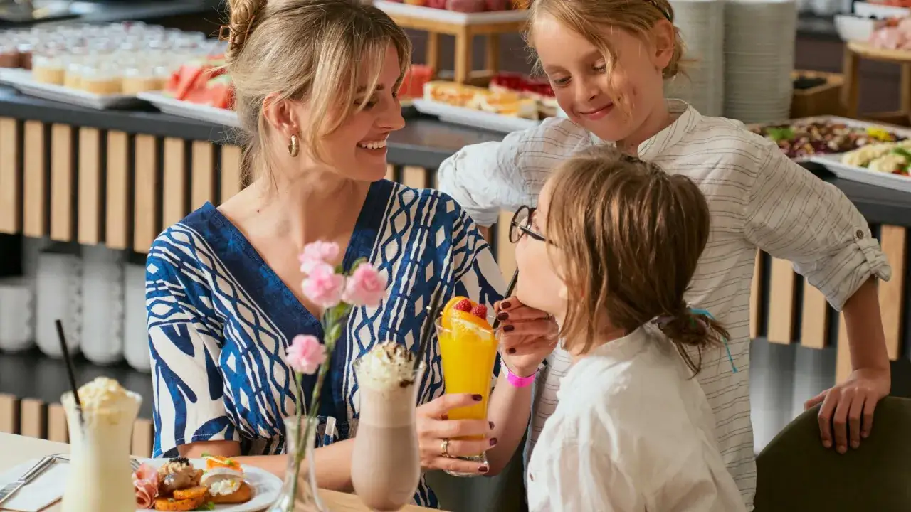 Rodzina cieszy się posiłkiem w restauracji z opcją all inclusive plus. Dzieci piją soki, a mama uśmiecha się do nich.