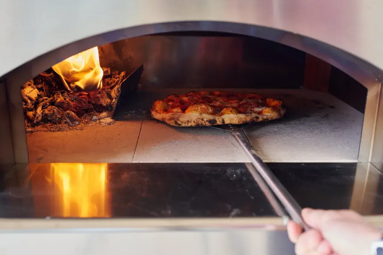 Una pizza masa madre dorada, lista para ser retirada del horno de leña, con brasas ardientes a un lado.
