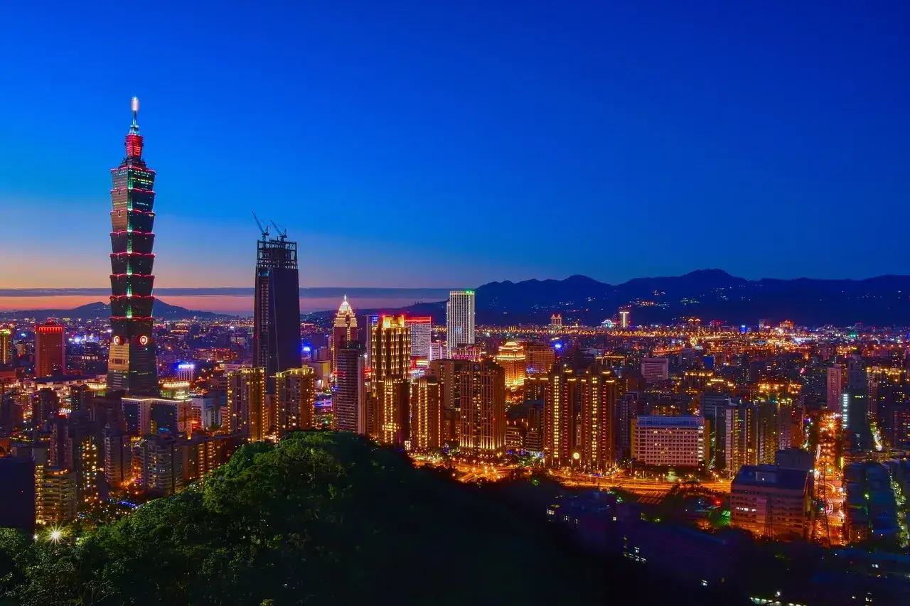 Wieczorny widok na Taipei, stolicę Tajwanu, z oświetlonymi drapaczami chmur i górami w tle.