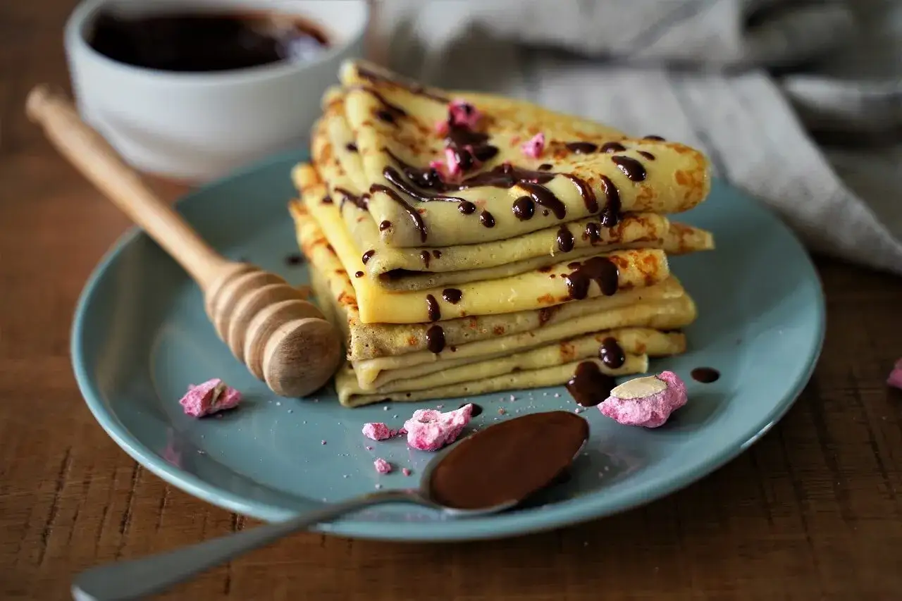 Pile de crêpes moelleuses arrosées de chocolat, décorées de quelques éclats roses. Une cuillère de chocolat et un mielier complètent cette gourmandise.