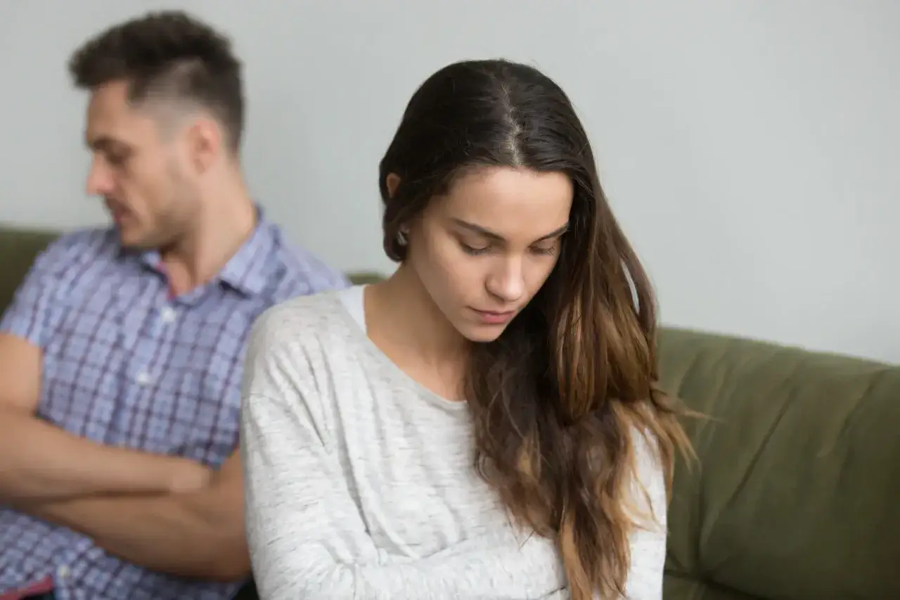Pareja sentada en un sof&aacute;, con la mujer mirando hacia abajo y el hombre de espaldas, reflejando el **apego intermitente pareja** y la distancia emocional.