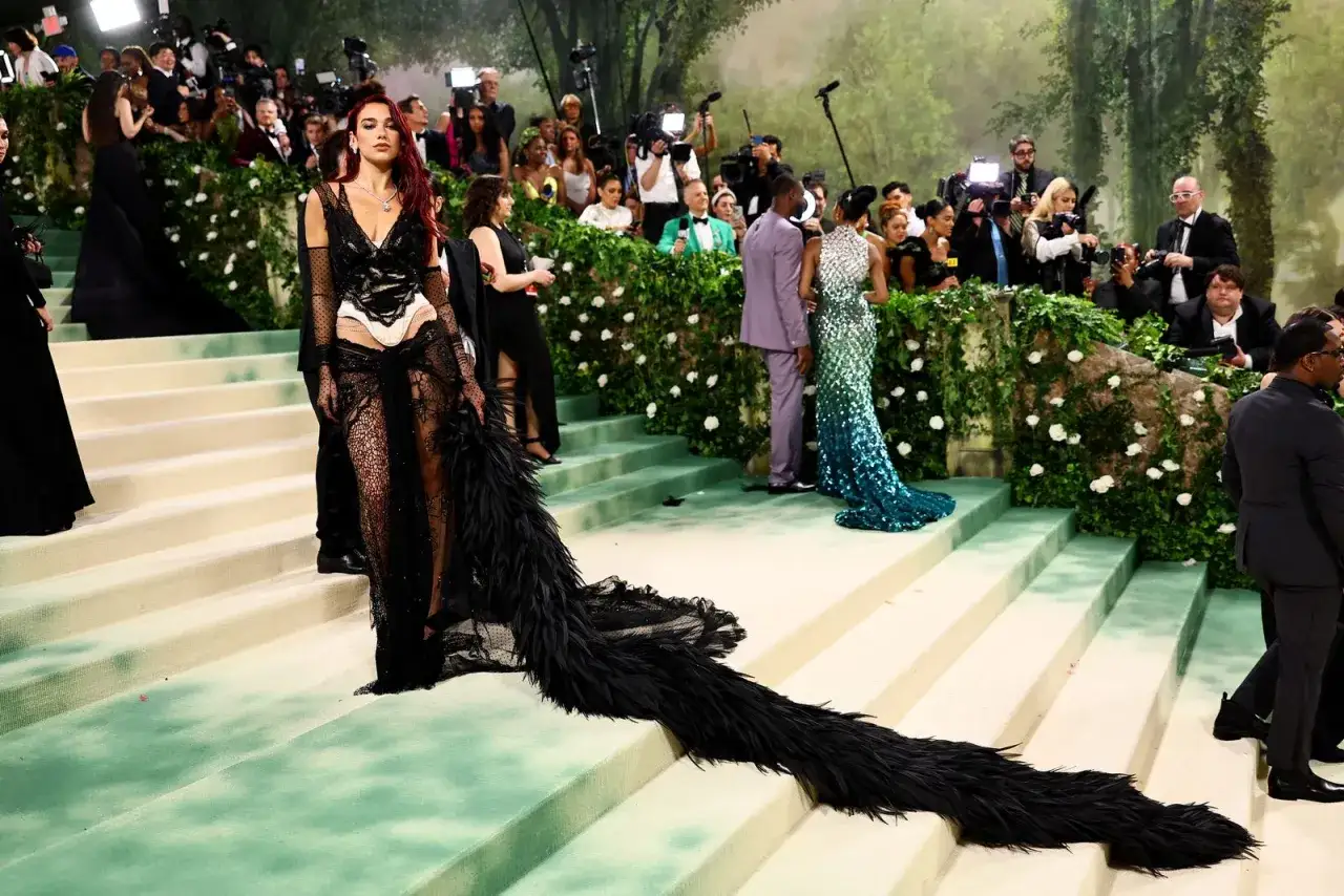 Dua Lipa makes a dramatic entrance at the Met Gala in a black lace gown with a dramatic feather train, captivating the cameras.