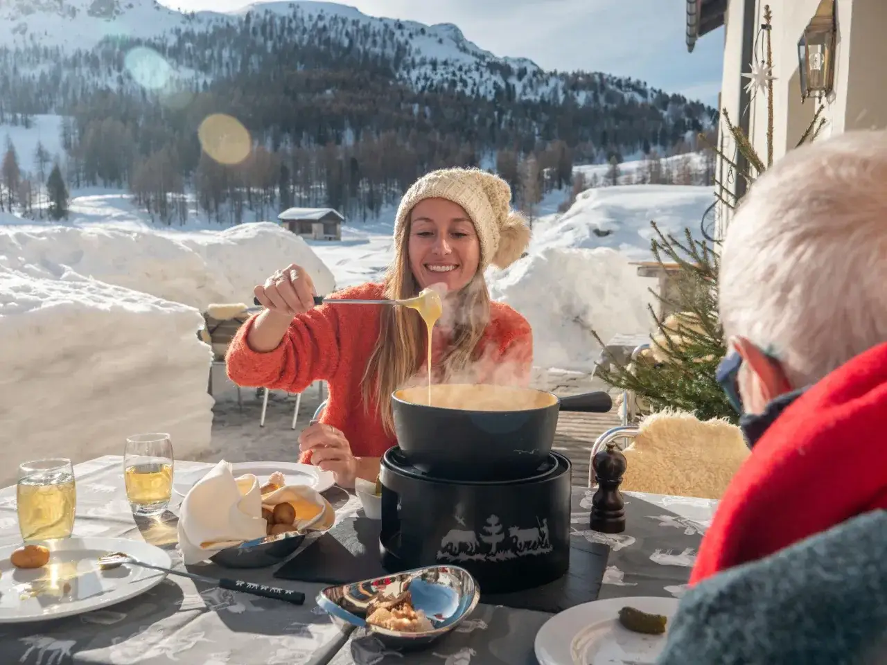 Une femme souriante déguste des fromages fondus suisses en terrasse enneigée, avec des montagnes en arrière-plan.