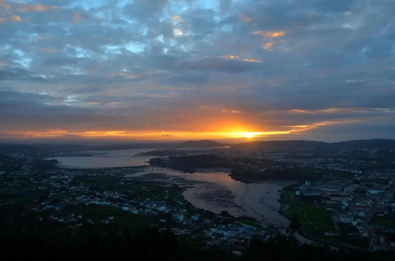 Vista desde un monte ventoso al amanecer, con el sol asomando entre las nubes y tiñendo el cielo de naranja y azul sobre una ciudad y un río.