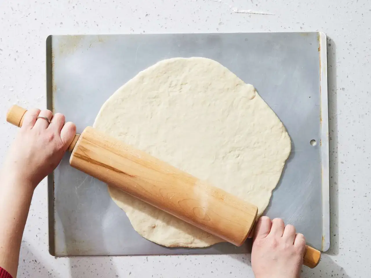 H&auml;nde rollen mit einem Holz-Nudelholz einen **schneller pizzateig ohne hefe** auf einem Backblech aus.