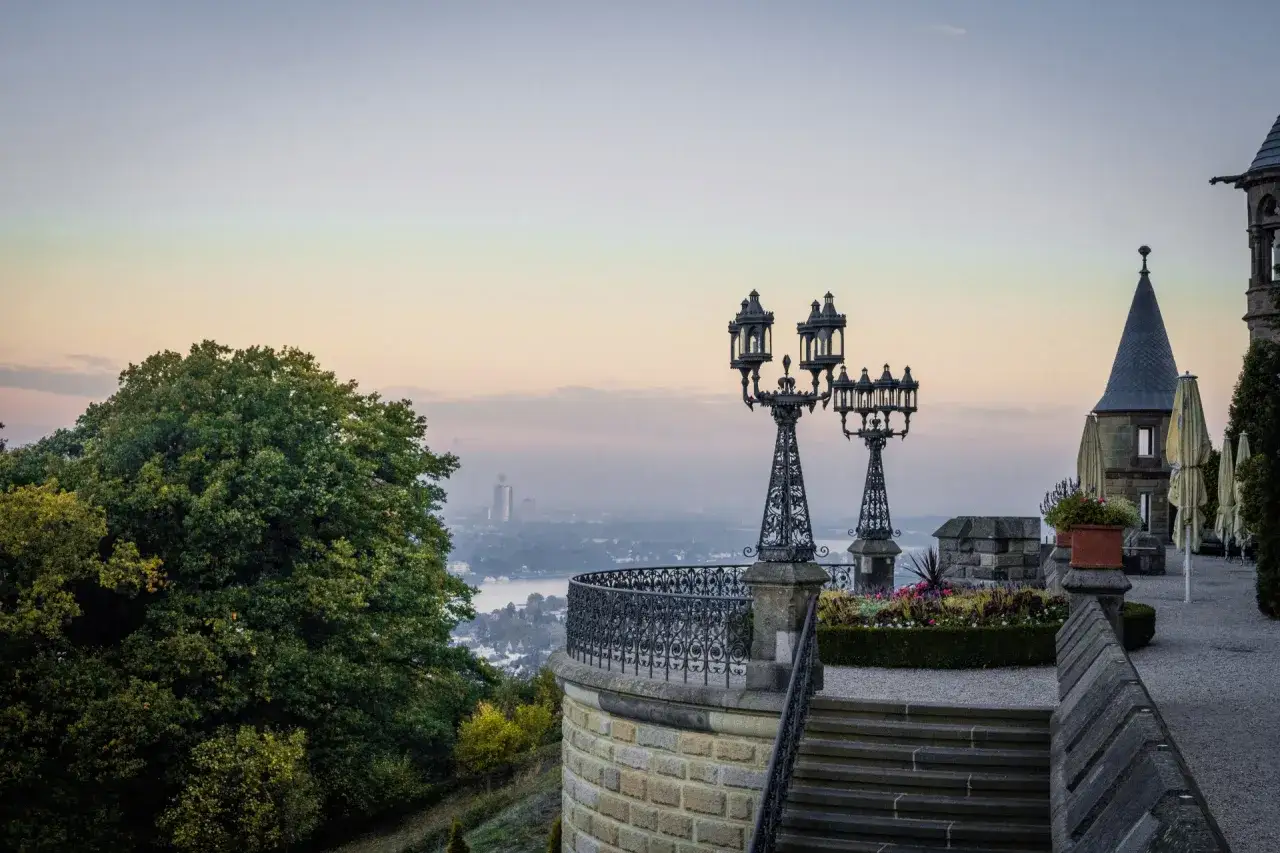 Blick vom Schloss Drachenburg auf eine neblige Stadtlandschaft mit Fluss. Zwei kunstvolle Laternen stehen auf einer Terrasse.