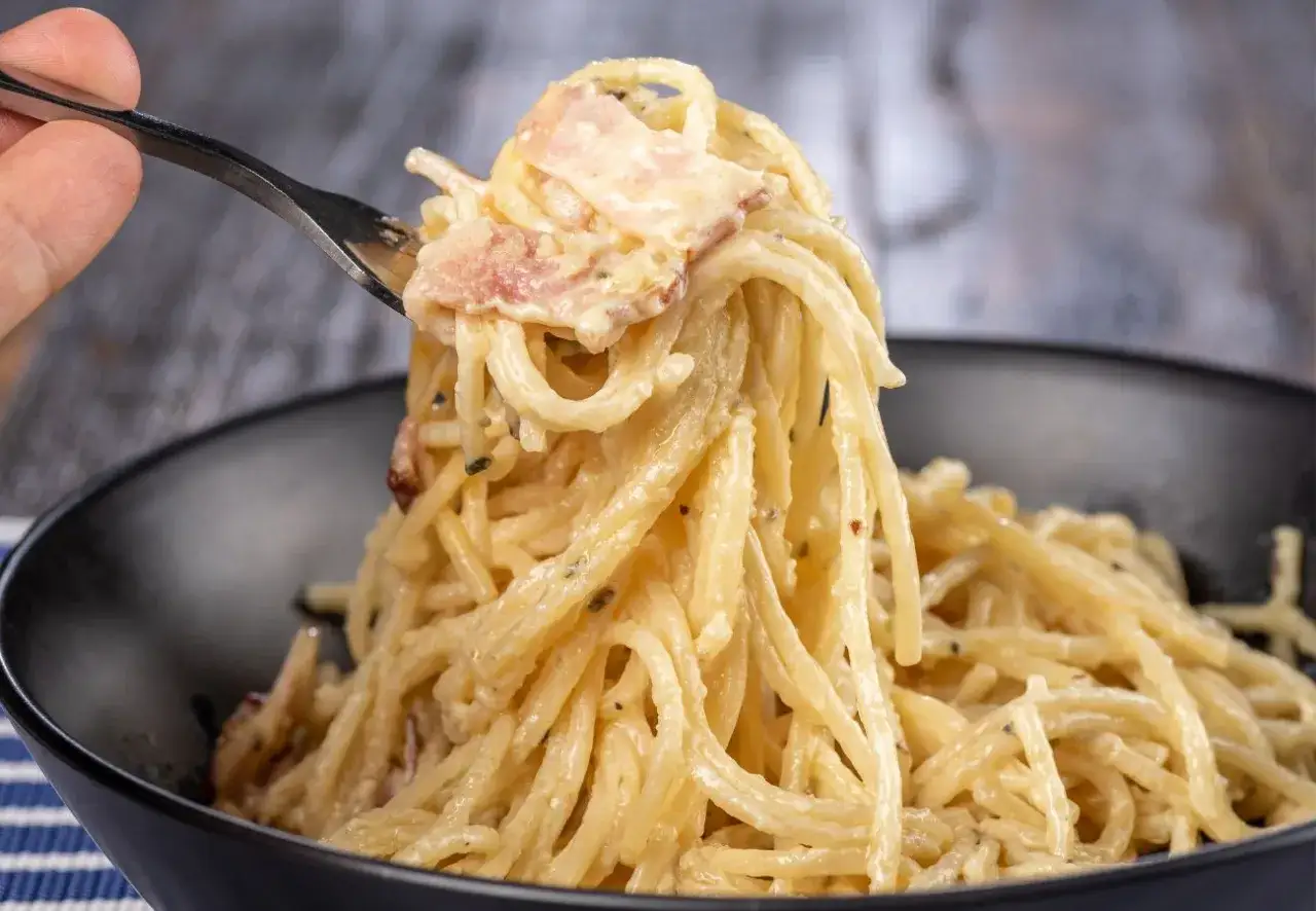 Un tenedor levanta un bocado cremoso de tallarines a la carbonara, con trozos de panceta y pimienta negra.