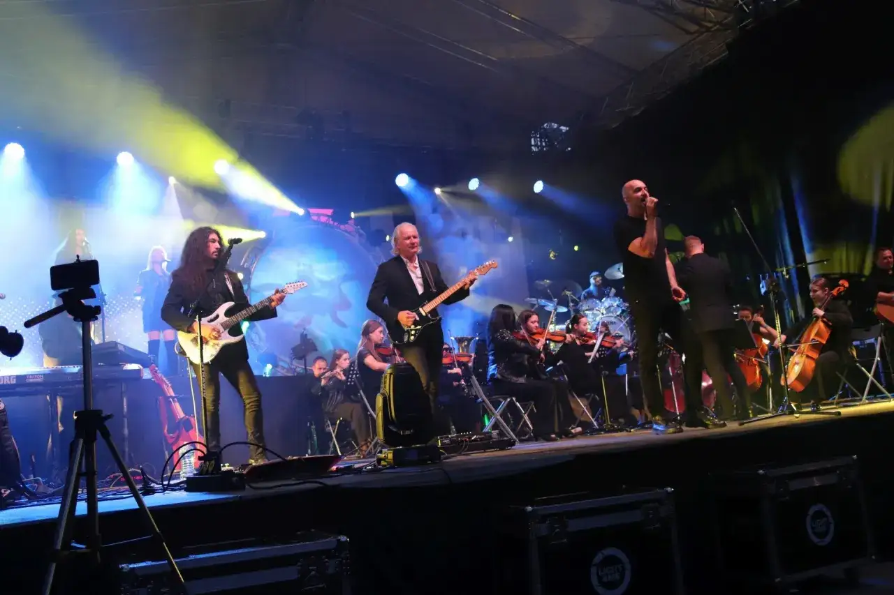 Ira i przyjaciele na scenie z orkiestrą. Muzycy grają na gitarach i instrumentach smyczkowych, wokalista śpiewa.