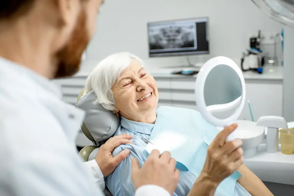 Uśmiechnięta starsza pani ogląda swoje zęby w lusterku, podczas gdy dentysta kładzie jej rękę na ramieniu. Nie martwi się już o zapach ryby z ust.