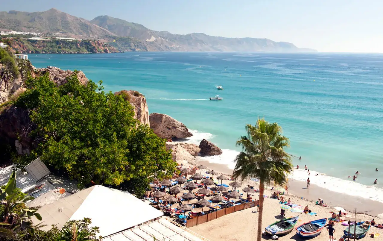 Costa del Sol w lutym: słoneczna plaża z palmami, turkusowe morze i g&oacute;ry w tle. Ludzie odpoczywają pod parasolami.