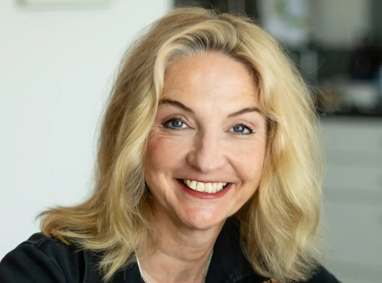 L&auml;chelnde Katja Reider mit blonden Haaren und blauen Augen, tr&auml;gt ein schwarzes Oberteil.