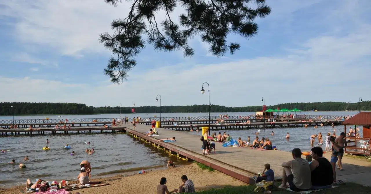 Ludzie odpoczywają i kąpią się nad jeziorem w Augustowie. Długie molo pełne plażowiczów i kąpiących się w wodzie.