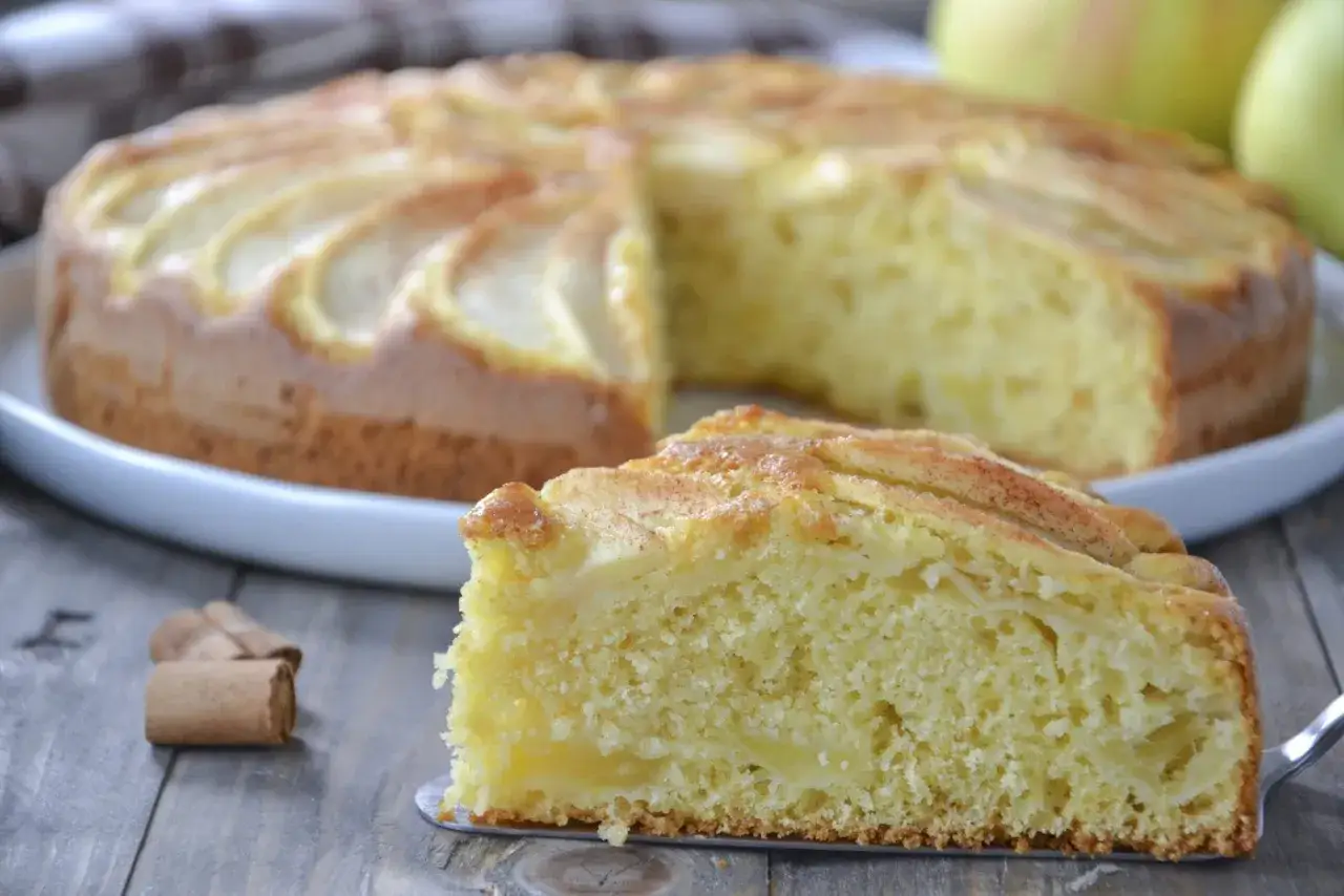 Una fetta di torta di mele senza burro, soffice e profumata, con mele a vista e cannella. La torta intera &egrave; sullo sfondo.