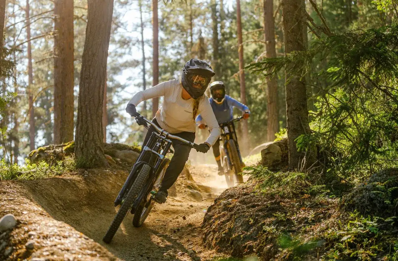 Tv&aring; personer cyklar utf&ouml;r en dammig stig i en solig skog.
