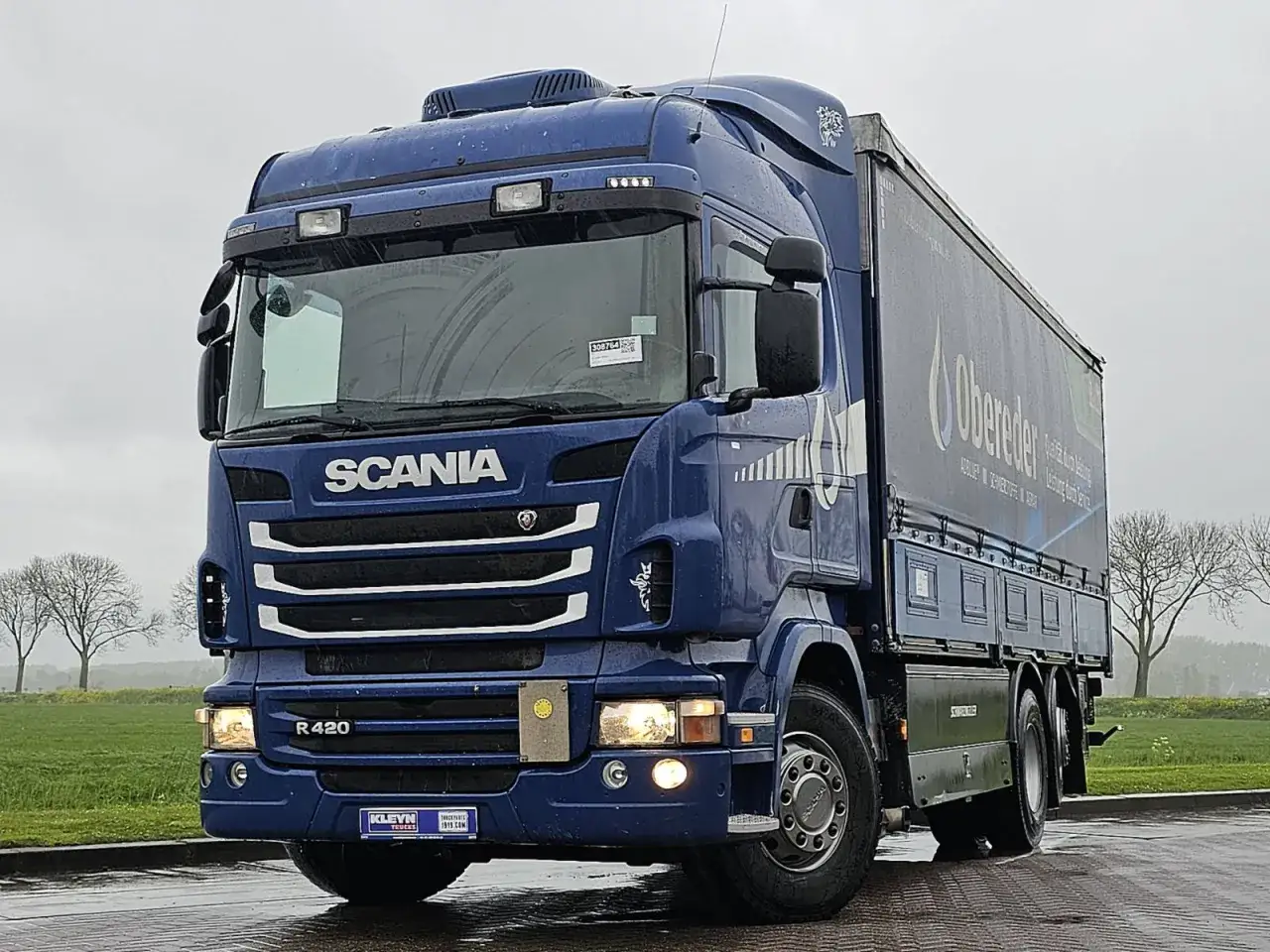 Niebieska Scania R420 z naczepą firmy Obereder stoi na mokrej drodze.