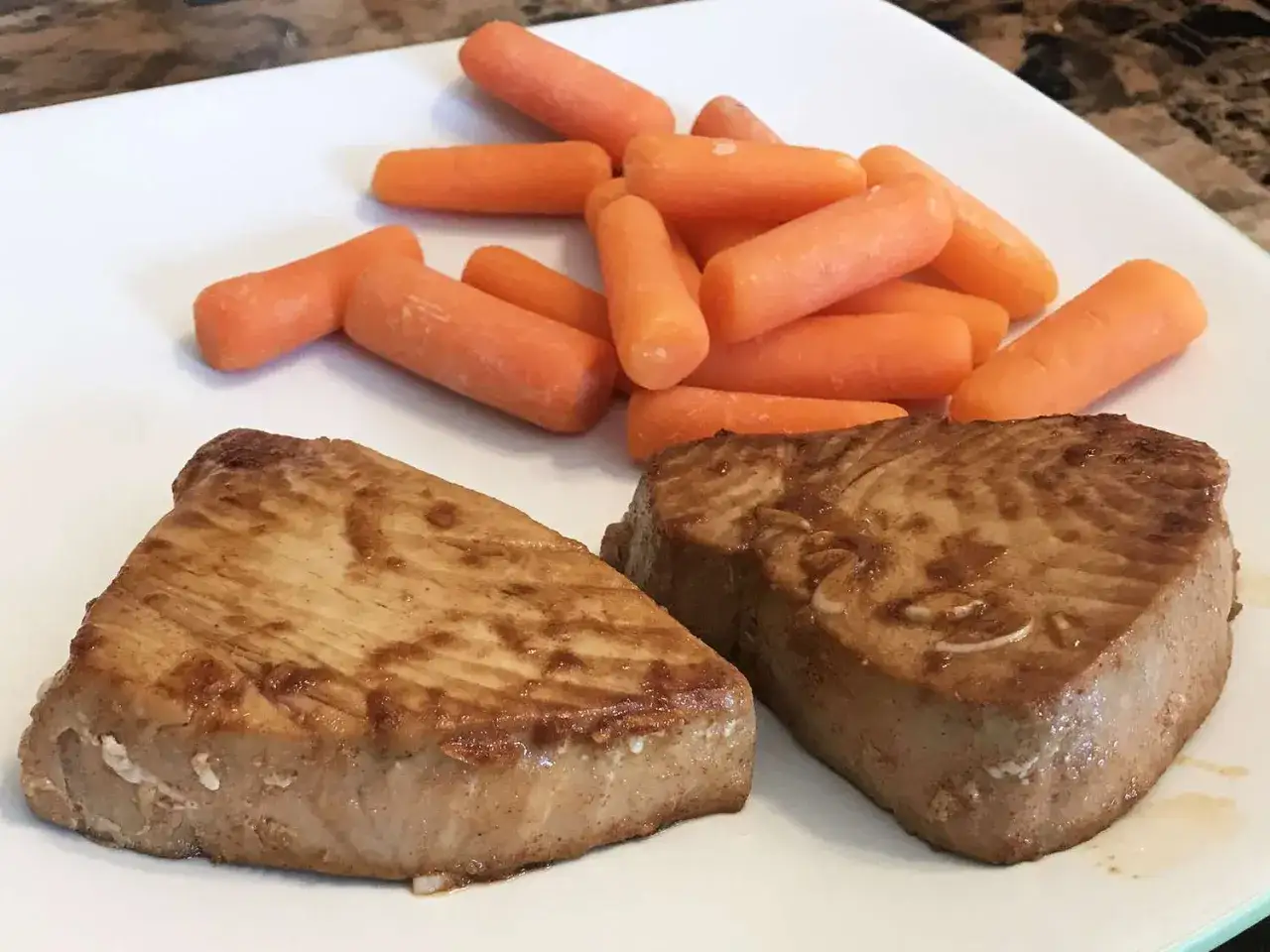 Zwei gebratene Thunfischsteaks mit einer Beilage aus Babykarotten auf einem weißen Teller.
