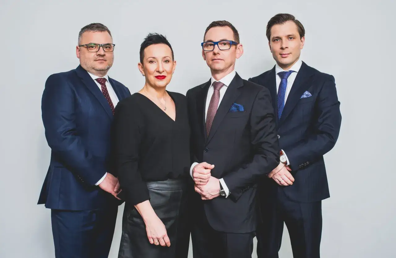 Cztery osoby w eleganckich garniturach i czarnej bluzce, stojące obok siebie na jasnym tle.