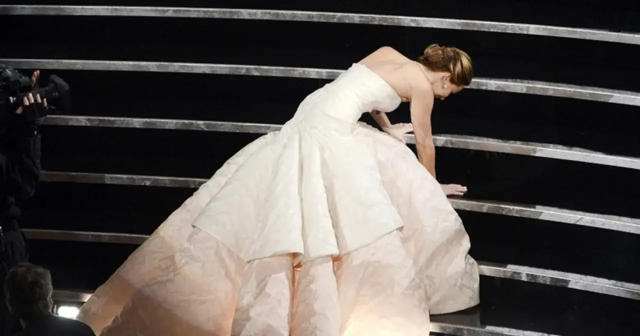 Jennifer Lawrence w białej sukni potyka się na schodach, przypominając scenę z jej filmów.