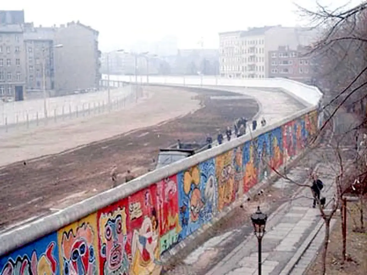 Kolorowy Mur Berliński, ozdobiony graffiti, tuż przed tym, kiedy upadł mur berliński. Ludzie spacerują po chodniku.