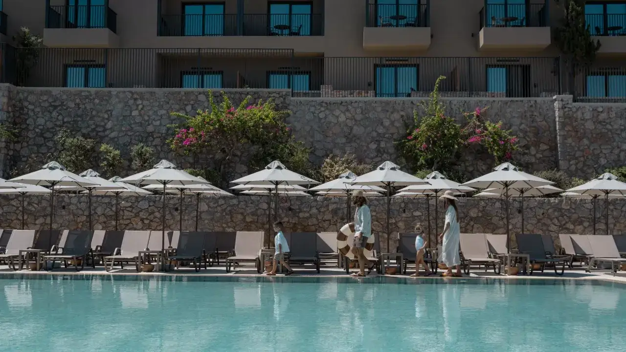 Familie spaziert am Pool entlang, mit Blick auf das Hotel am Elios Hill, Kreta. Sonnenschirme und Liegen laden zum Entspannen ein.