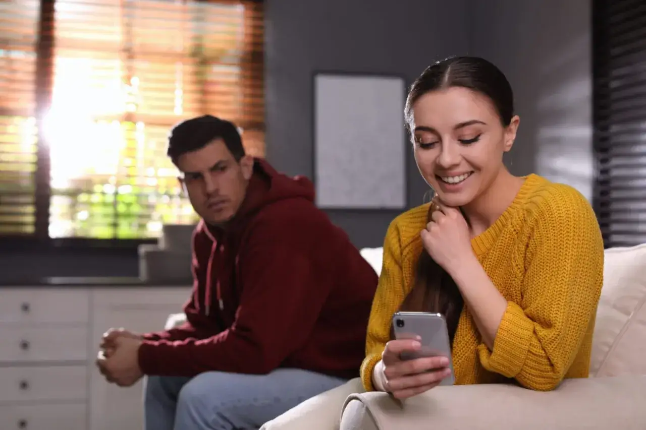 Mężczyzna z zazdrością patrzy na kobietę, która śmieje się, przeglądając telefon. Siedzą obok siebie, ale ona jest pochłonięta wirtualnym światem, a on czuje się pominięty, jakby był na ławce rezerwowych w ich związku.