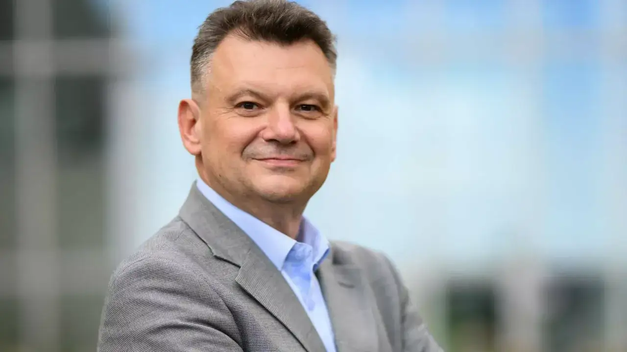 Uśmiechnięty mężczyzna, january ciszewski, w szarym garniturze i niebieskiej koszuli, na tle rozmazanego budynku.