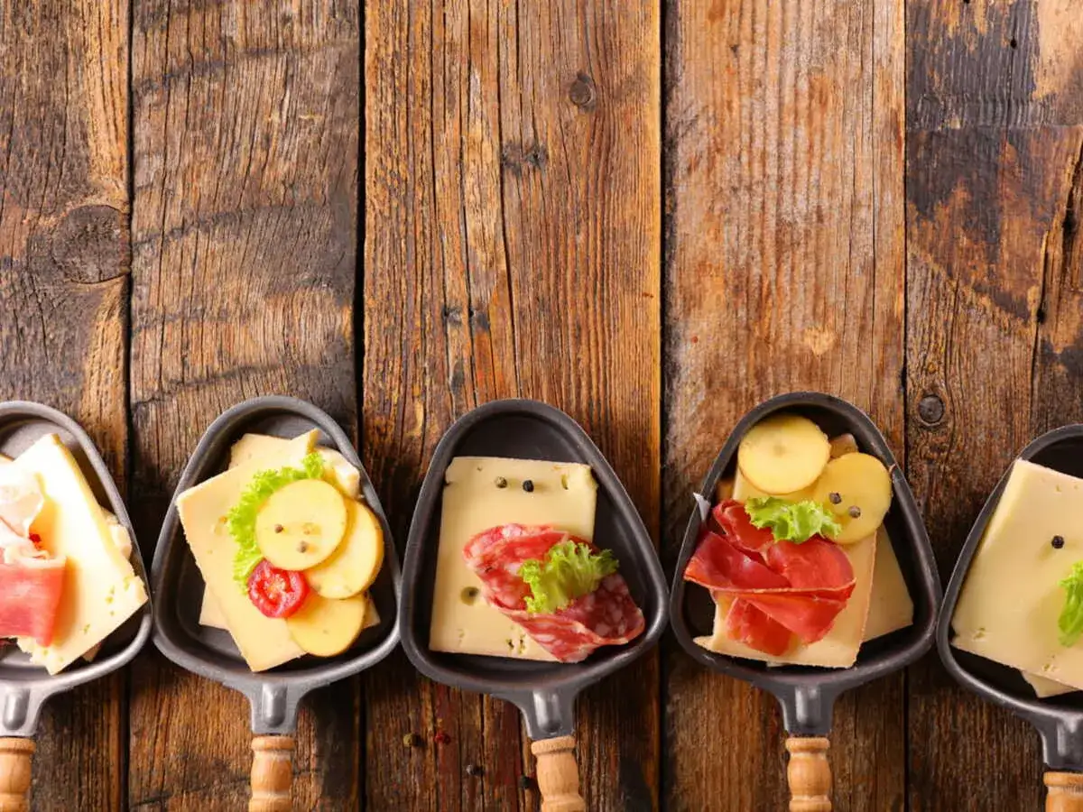 Des coupelles pour un apéritif raclette, garnies de fromage, charcuterie et pommes de terre, sur un fond de bois rustique.
