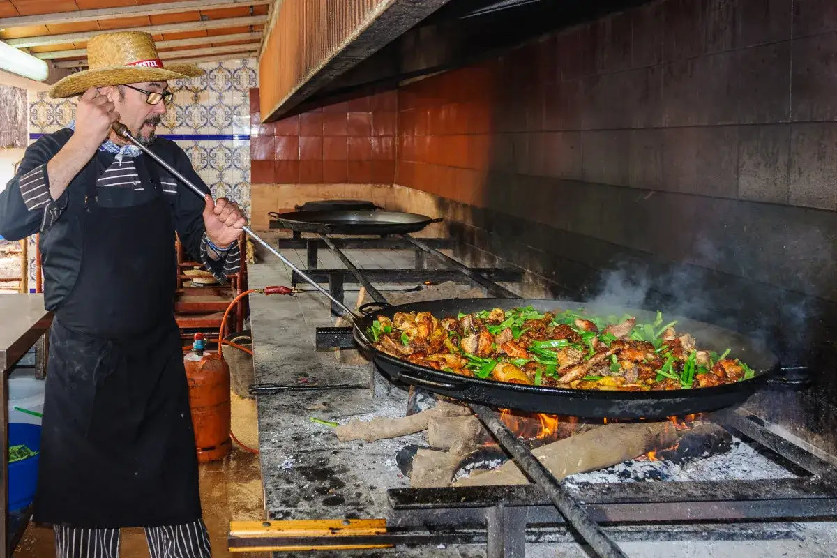 Chef con sombrero de paja prepara la mejor paella en Valencia, cocinada a le&ntilde;a en una gran paellera humeante.