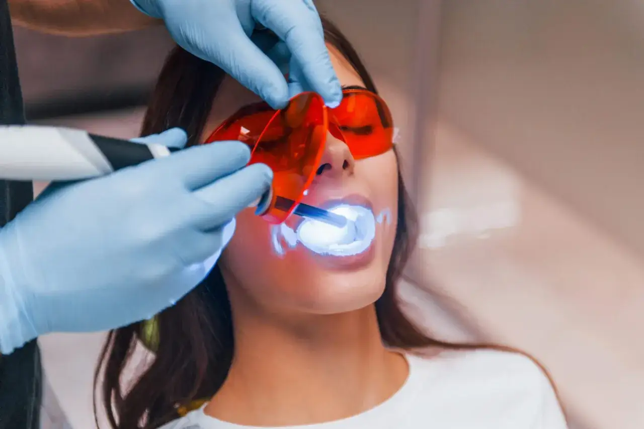 Dentysta w rękawiczkach naświetla zęby pacjentki lampą, co jest częścią zabiegu. Czy wybielanie zębów jest zdrowe? Zapytaj specjalistę.