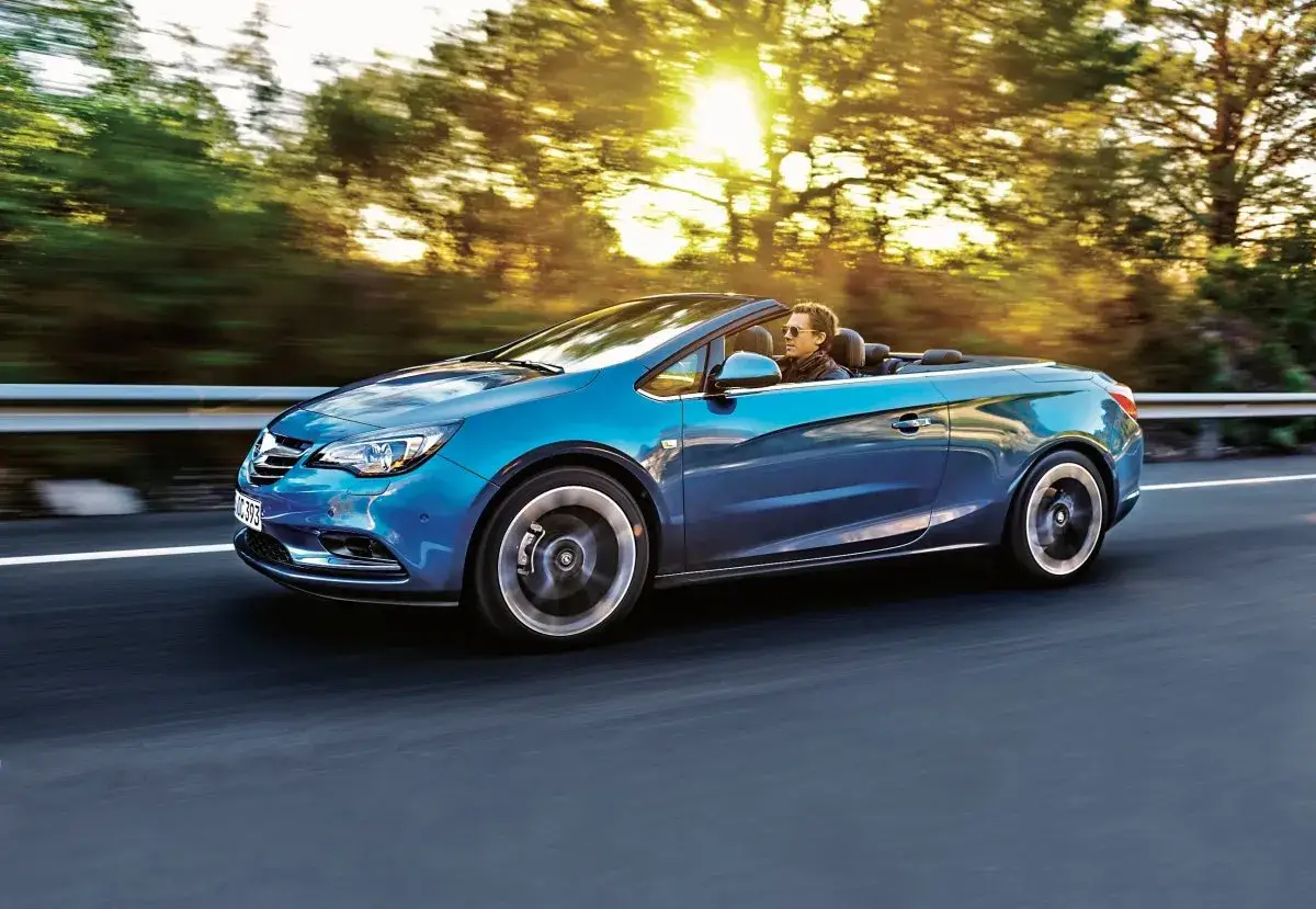 Błękitny Opel Cascada z opuszczonym dachem sunie po drodze, a słońce przebija się przez drzewa.