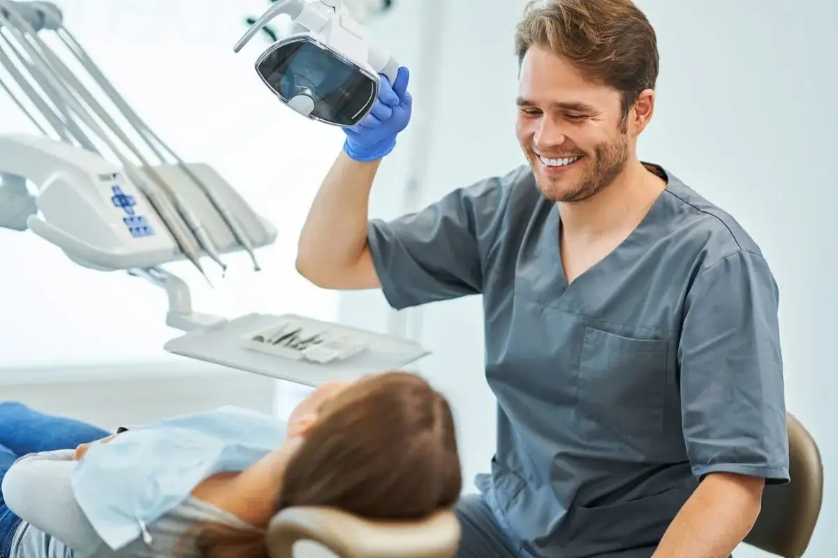 Uśmiechnięty dentysta w niebieskiej rękawiczce reguluje lampę, by lepiej widzieć co widzi dentysta. Pacjentka leży na fotelu.