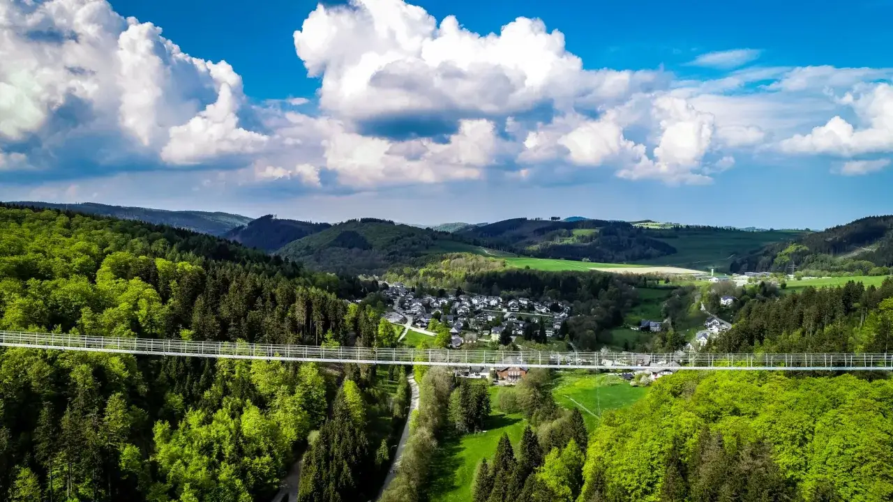 Der Skywalk Willingen &uuml;berspannt ein gr&uuml;nes Tal mit W&auml;ldern und einem Dorf. Wolken ziehen &uuml;ber den blauen Himmel.