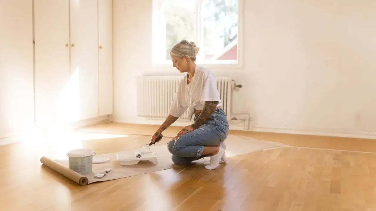 Une femme tatoueuse, en jean déchiré, s'agenouille pour peindre un parquet stratifié. Matériel de bricolage à portée de main.