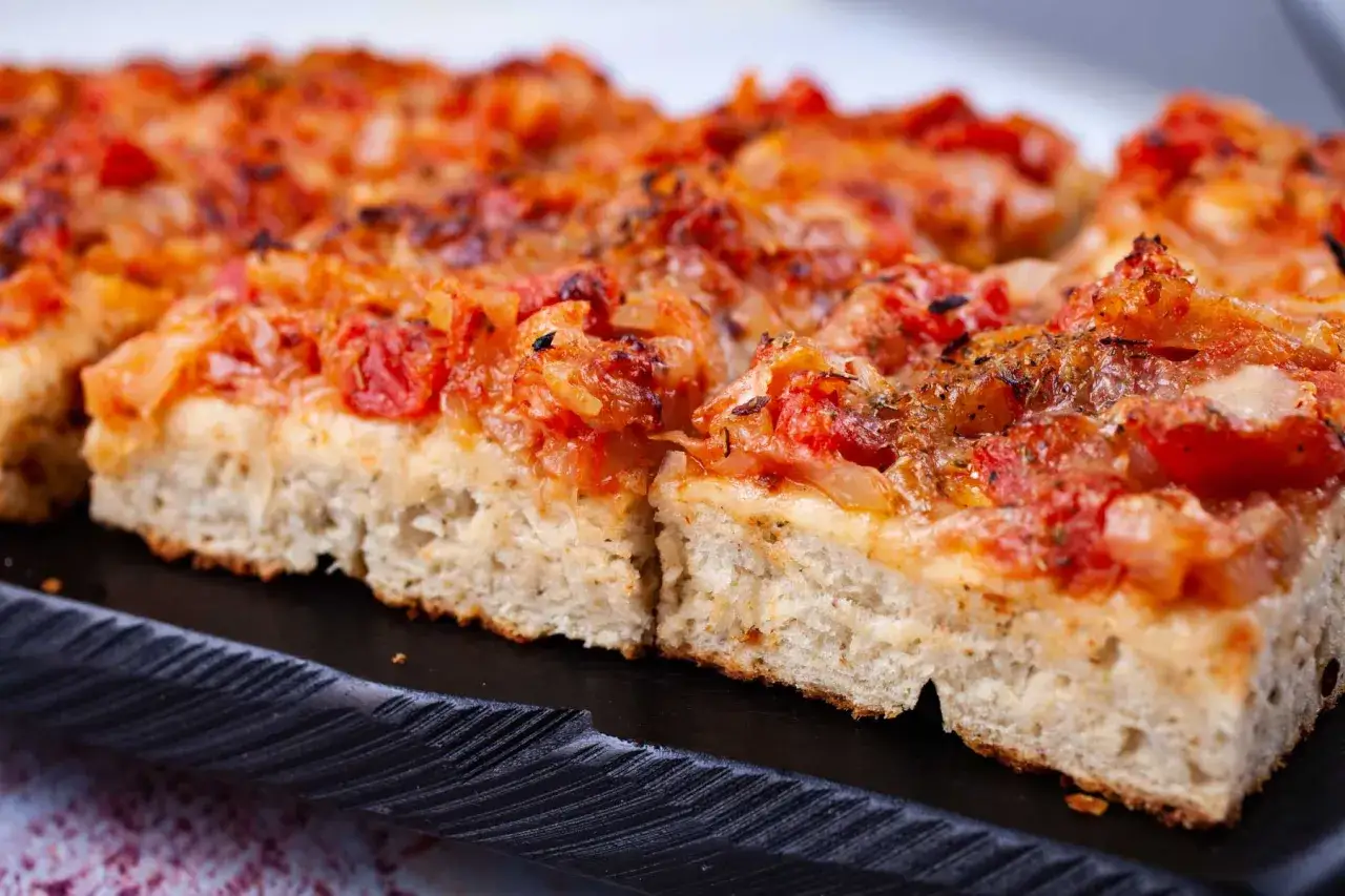 En bit av en tjock, fluffig pizza siciliana med tomater och lök, serverad på en svart platta.