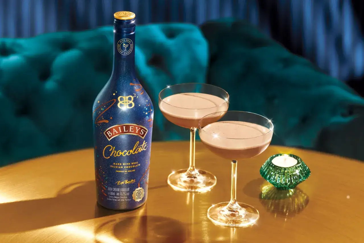 Baileys co to? Butelka Baileys Chocolate i dwa kieliszki z tym likierem na złotym stole, w tle aksamitna kanapa.