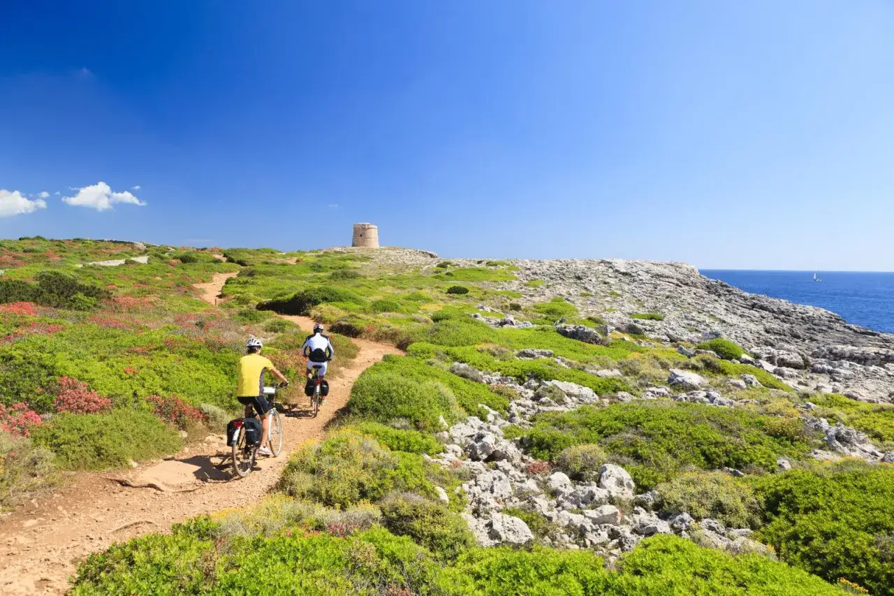 Ciclistas disfrutan del carril bici en Menorca, rodeados de vegetaci&oacute;n y con una torre vig&iacute;a al fondo.