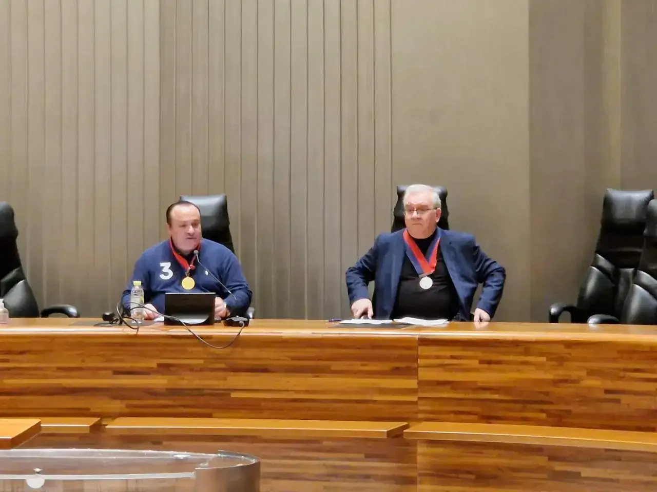 Deux hommes, dont un juge départiteur, sont assis à une table en bois. L'un porte un pull bleu avec un