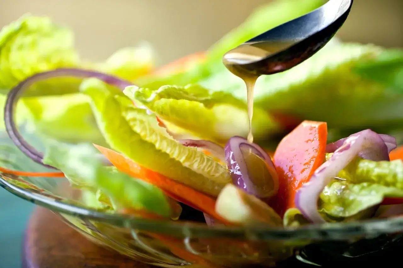 Una cuchara vierte vinagreta de mostaza sobre una ensalada fresca de lechuga, zanahoria y cebolla morada en un bol de cristal.