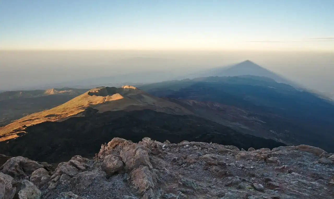 Widok z góry na wulkaniczny krajobraz Teneryfy. W oddali widać cień góry, a na pierwszym planie skaliste zbocze. Wejście na Teide to niezapomniane przeżycie.