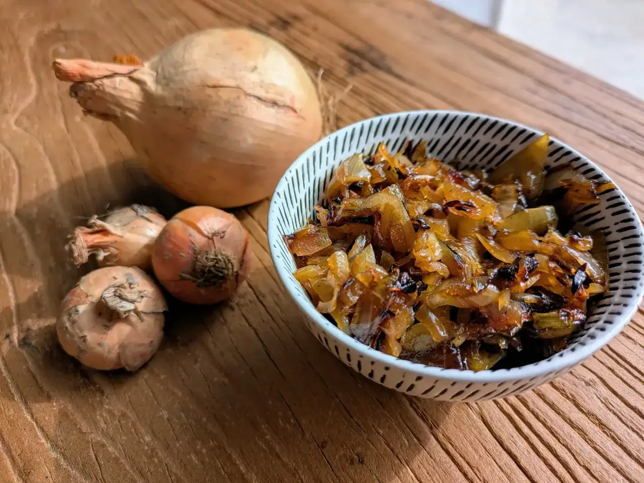 Un bol de oignons caramélisés dorés et tendres, à côté d'oignons frais sur une table en bois.