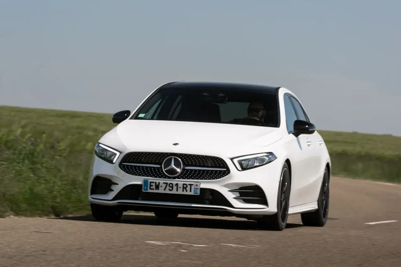 Une Mercedes-Benz Classe A blanche roule sur une route de campagne. Le conducteur porte des lunettes de soleil.