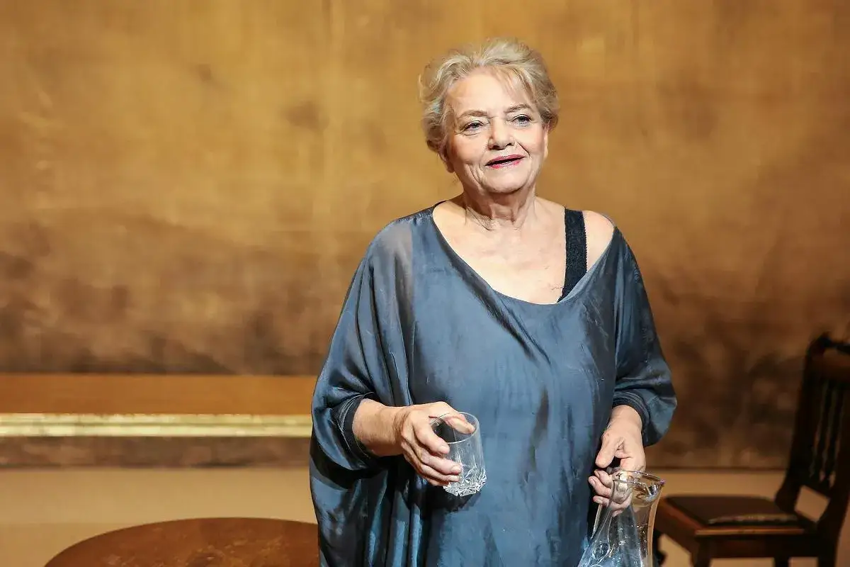 Anna Seniuk obecnie, w luźnej, szarej sukni, trzyma szklankę i karafkę.