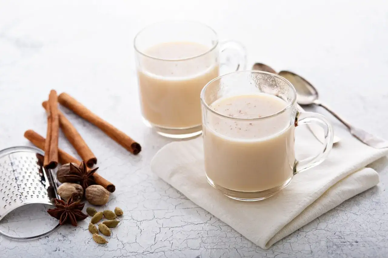 Dwa kubki aromatycznego napoju, przypominającego chai. Obok cynamon, kardamon, anyż i gałka muszkatołowa.