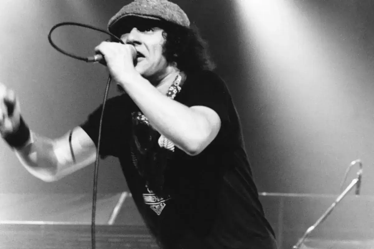 Młody Brian Johnson w czapce i z mikrofonem, w pełnym energii występie na scenie.