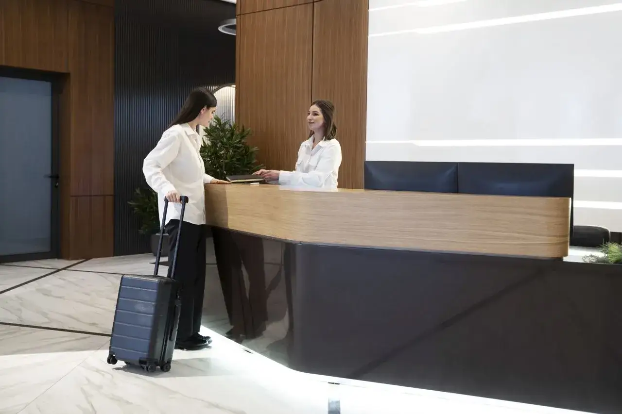 Kobieta z walizką przy recepcji hotelowej. Dowiaduje się, od ilu lat można zarezerwować hotel.