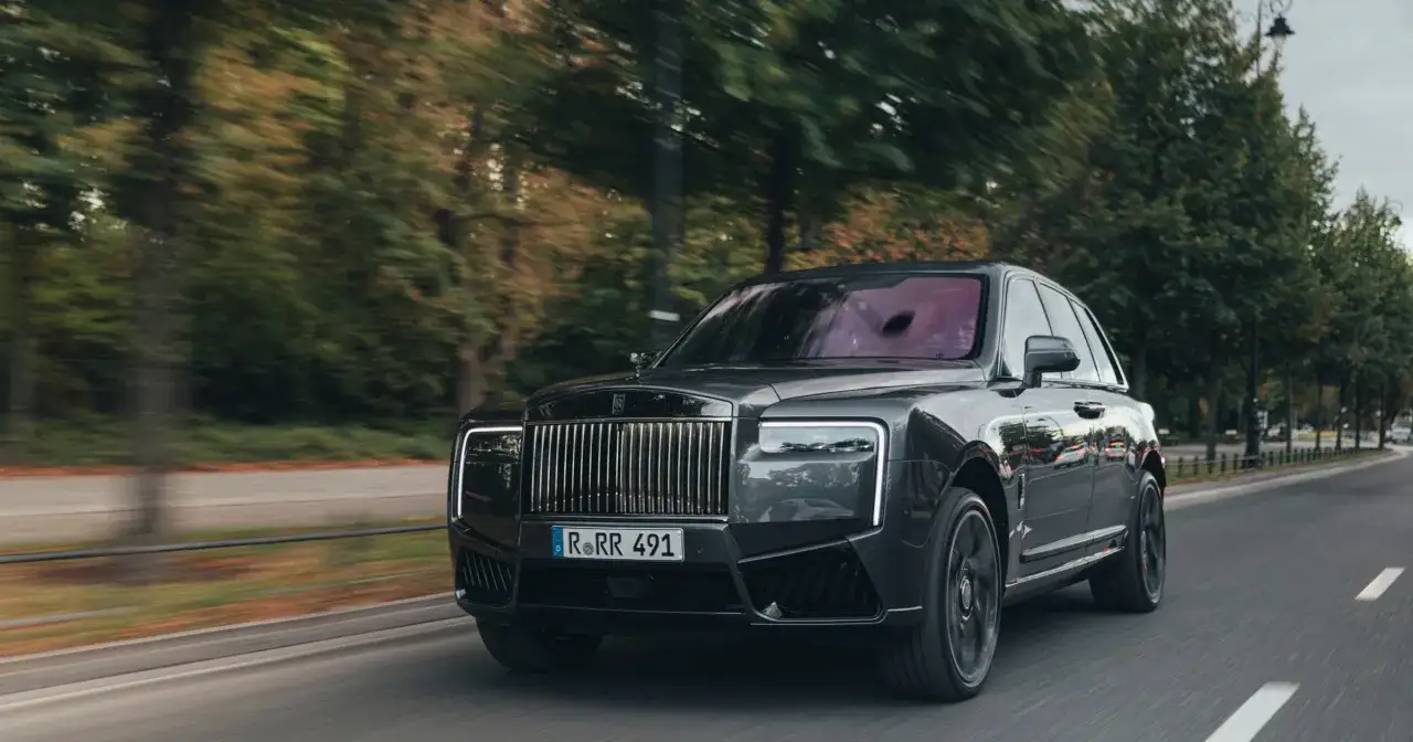 Czarny Rolls-Royce Cullinan, prawdopodobnie najdroższe auto w Polsce, pędzi po ulicy wśród drzew.