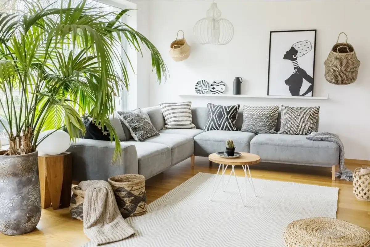 Sof&aacute; gris claro con cojines de patrones geom&eacute;tricos y una planta grande. Los colores que combinan con el gris claro son el blanco, negro y tonos tierra, creando un ambiente moderno y acogedor.